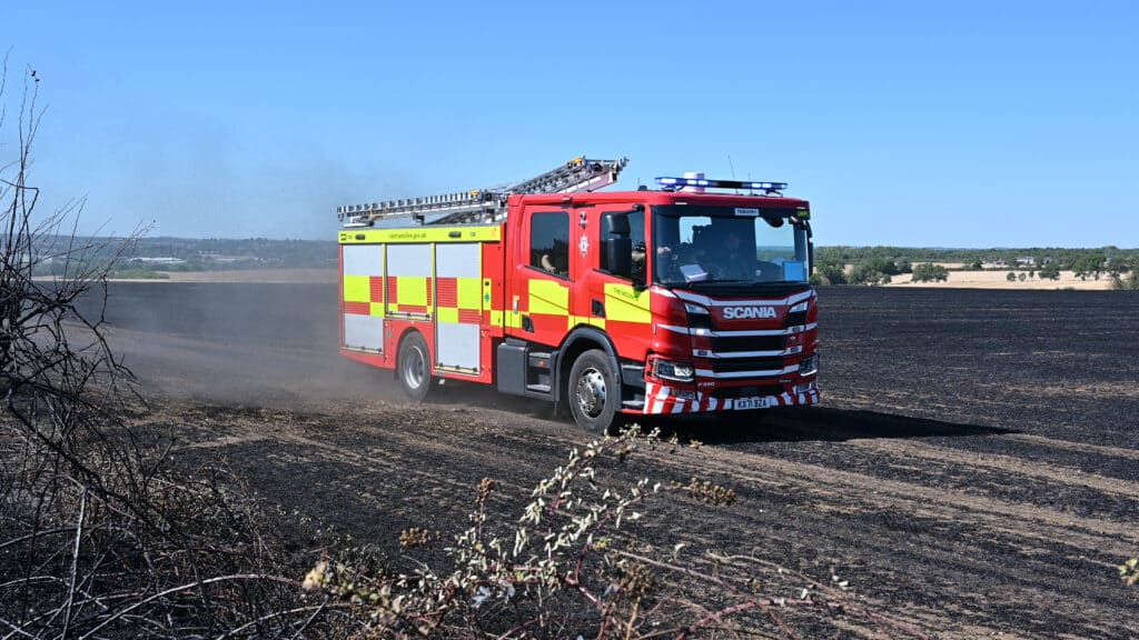 Two red Northamptonshire Fire and Rescue Service fire engines drive across a dry harvested field, raising dust behind them. The lead vehicle has blue lights illuminated, with a second fire engine following in the background. Trees line the edge of the field under a bright, clear sky.