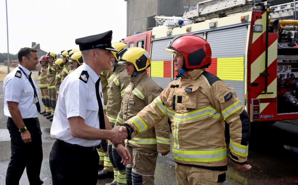 Firefighter recruits take part in a ladder training exercise beside a multi-storey concrete training tower at the Fire Service College. Several firefighters in full protective clothing work together to raise and secure an extension ladder to an upper-level walkway, while others kneel nearby ready to support the operation. A fire engine is parked alongside the training ground, and a person stands on the upper platform as part of the exercise. The scene takes place outdoors in daylight.