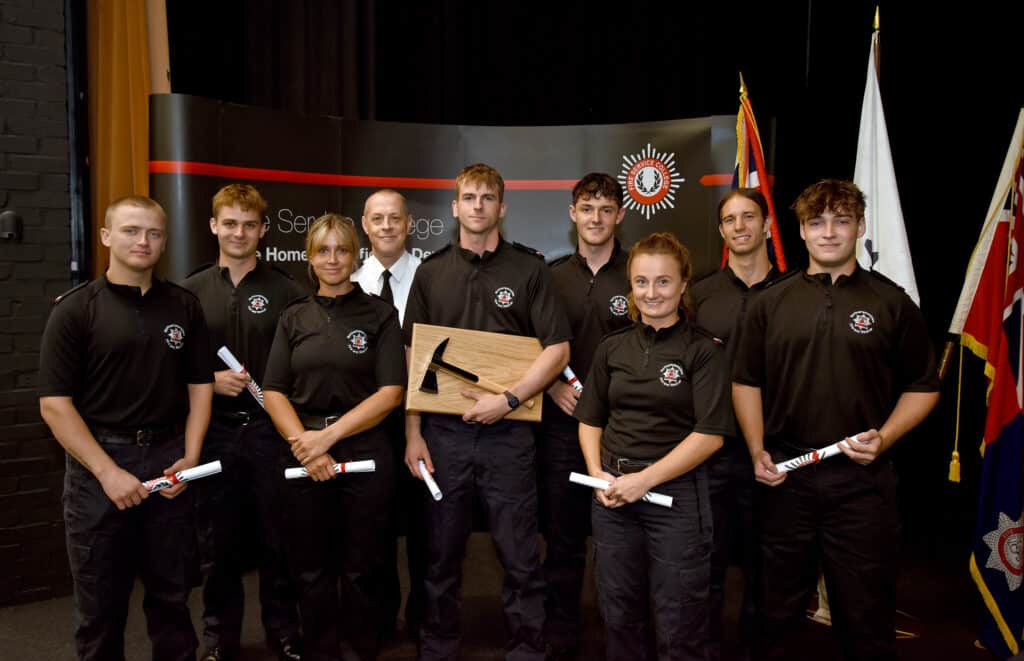 Group photo of eight firefighter recruits and one instructor standing on a stage at a graduation ceremony. The recruits wear black uniform polo shirts and hold rolled certificates. One recruit in the centre holds a mounted ceremonial axe award. Behind the group is a Fire Service College backdrop and several flags, including Union flags. The photograph is taken indoors under stage lighting. Provide your feedback on BizChat Make this alt text shorterRewrite it for screen readersCreate a more detailed image description