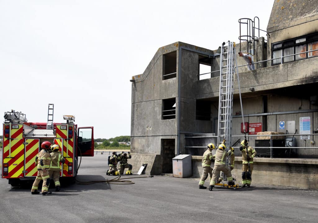 Firefighter recruits take part in a ladder training exercise beside a multi-storey concrete training tower at the Fire Service College. Several firefighters in full protective clothing work together to raise and secure an extension ladder to an upper-level walkway, while others kneel nearby ready to support the operation. A fire engine is parked alongside the training ground, and a person stands on the upper platform as part of the exercise. The scene takes place outdoors in daylight.